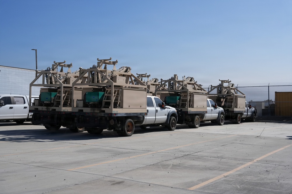 Multiple white flatbed pickup trucks are parked in a paved lot, each carrying identical tan/desert-colored military or surveillance equipment mounted on their beds. The equipment appears to be FLIR-branded sensor or surveillance systems with structural frames and mast assemblies. The setting appears to be a secure facility or staging area based on the fencing visible in the background.