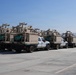 Multiple white flatbed pickup trucks are parked in a paved lot, each carrying identical tan/desert-colored military or surveillance equipment mounted on their beds. The equipment appears to be FLIR-branded sensor or surveillance systems with structural frames and mast assemblies. The setting appears to be a secure facility or staging area based on the fencing visible in the background.