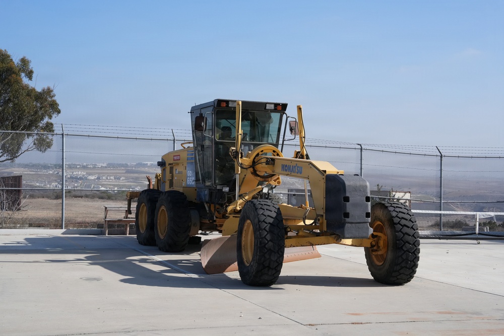 A yellow Komatsu motor grader is parked on a concrete surface behind a chain-link fence topped with barbed wire. The heavy construction equipment appears stationary, with its front blade lowered near the ground. The background reveals an open landscape with distant residential development and a clear blue sky.