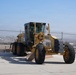A yellow Komatsu motor grader is parked on a concrete surface behind a chain-link fence topped with barbed wire. The heavy construction equipment appears stationary, with its front blade lowered near the ground. The background reveals an open landscape with distant residential development and a clear blue sky.