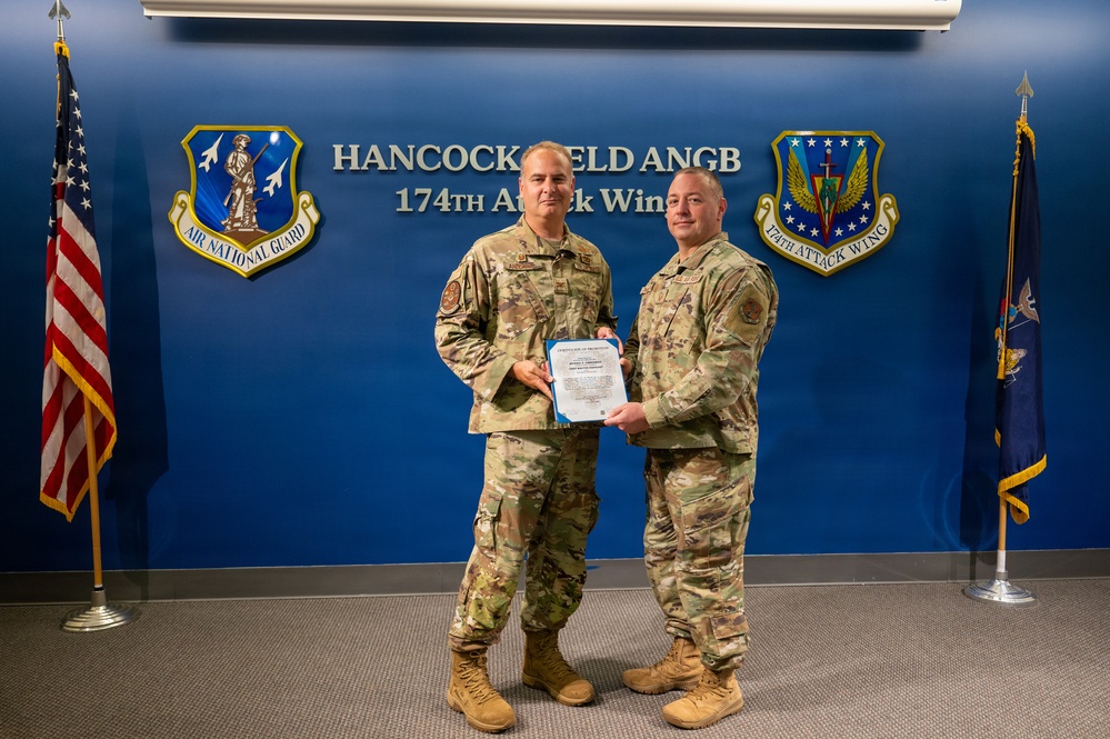 Two military personnel in OCP (Operational Camouflage Pattern) uniforms pose together holding a certificate or official document at Hancock Field Air National Guard Base, home of the 174th Attack Wing. The setting appears to be an official ceremony, likely a promotion or award presentation. The backdrop features the Air National Guard and 174th Attack Wing insignia on a blue wall.