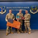 Three military personnel in OCP (Operational Camouflage Pattern) uniforms pose for a photo at Hancock Field Air National Guard Base, home of the 174th Attack Wing. They are holding what appear to be handcrafted wooden American flag art pieces and a decorative metal eagle/flag plaque, likely presented as gifts or awards. The setting is a formal backdrop wall displaying unit insignia and signage.