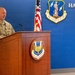 A U.S. Air Force military officer in OCP (Operational Camouflage Pattern) uniform is speaking at a wooden podium bearing the 174th Attack Wing emblem. The setting is an official military venue at Hancock Field, home of the 174th Attack Wing, Air National Guard. An American flag is visible in the background alongside the Air National Guard shield insignia on a blue wall.