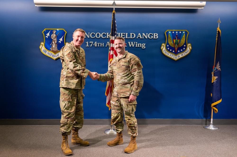 Two military personnel in OCP (Operational Camouflage Pattern) uniforms are shaking hands in front of a blue wall displaying the Hancock Field ANGB (Air National Guard Base) 174th Attack Wing signage. The setting appears to be an official ceremony or recognition event at the Air National Guard installation. Both individuals are smiling, suggesting a positive or celebratory occasion.