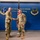 Murray Shakes Durant’s Hand During Pinning Ceremony Two military personnel in OCP (Operational Camouflage Pattern) uniforms are shaking hands in front of a blue wall displaying the Hancock Field ANGB (Air National Guard Base) 174th Attack Wing signage. The setting appears to be an official ceremony or recognition event at the Air National Guard installation. Both individuals are smiling, suggesting a positive or celebratory occasion.