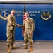 Two military personnel in OCP (Operational Camouflage Pattern) uniforms are shaking hands in front of a blue wall displaying the Hancock Field ANGB (Air National Guard Base) 174th Attack Wing signage. The setting appears to be an official ceremony or recognition event at the Air National Guard installation. Both individuals are smiling, suggesting a positive or celebratory occasion.