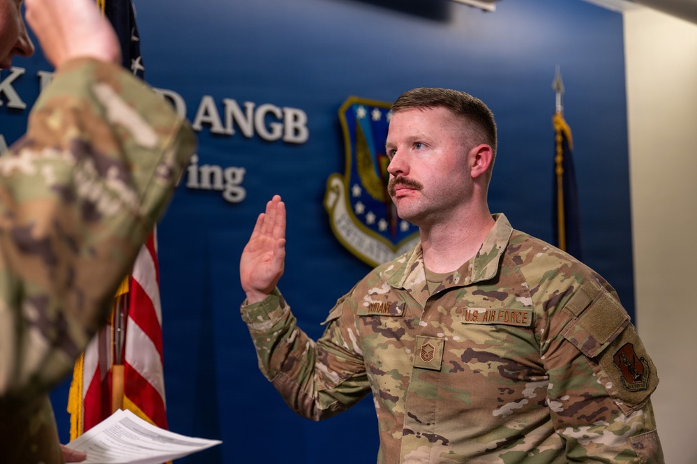A U.S. Air Force senior non-commissioned officer is taking an oath of office or reenlistment ceremony at what appears to be an Air National Guard Base (ANGB). The service member, whose name tag reads 'DURANT,' is raising his right hand while facing an officiating officer. The setting includes official military insignia, an Air Force shield emblem, and an American flag.