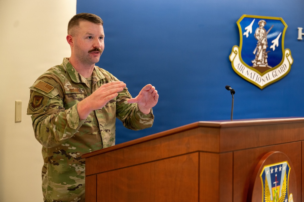 A U.S. Air National Guard service member in OCP (Operational Camouflage Pattern) uniform is speaking at a wooden podium. He is gesturing with both hands while addressing an audience in what appears to be an official briefing or ceremony room. The Air National Guard emblem is prominently displayed on the blue wall behind him.