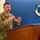 Durant Addresses Crowd During Pinning Ceremony A U.S. Air National Guard service member in OCP (Operational Camouflage Pattern) uniform is speaking at a wooden podium. He is gesturing with both hands while addressing an audience in what appears to be an official briefing or ceremony room. The Air National Guard emblem is prominently displayed on the blue wall behind him.
