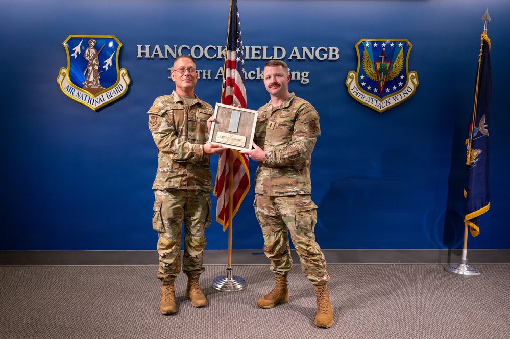 Two military personnel in OCP (Operational Camouflage Pattern) uniforms are posing together at Hancock Field Air National Guard Base, home of the 174th Attack Wing. They are jointly holding what appears to be a wooden plaque or award bearing the name 'Lucas Durant' with a rank of Lieutenant. The setting is an official ceremonial backdrop with unit insignia and flags.