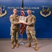 Two military personnel in OCP (Operational Camouflage Pattern) uniforms are posing together at Hancock Field Air National Guard Base, home of the 174th Attack Wing. They are jointly holding what appears to be a wooden plaque or award bearing the name 'Lucas Durant' with a rank of Lieutenant. The setting is an official ceremonial backdrop with unit insignia and flags.