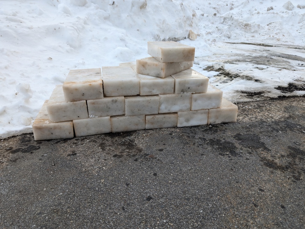 A pyramid-shaped stack of rectangular white/cream-colored blocks is arranged on a wet asphalt surface. The blocks appear to be handmade soap bars, stacked in a stepped pyramid formation with approximately 3-4 rows. The setting is outdoors in winter conditions, with snow visible in the background.