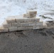 A pyramid-shaped stack of rectangular white/cream-colored blocks is arranged on a wet asphalt surface. The blocks appear to be handmade soap bars, stacked in a stepped pyramid formation with approximately 3-4 rows. The setting is outdoors in winter conditions, with snow visible in the background.