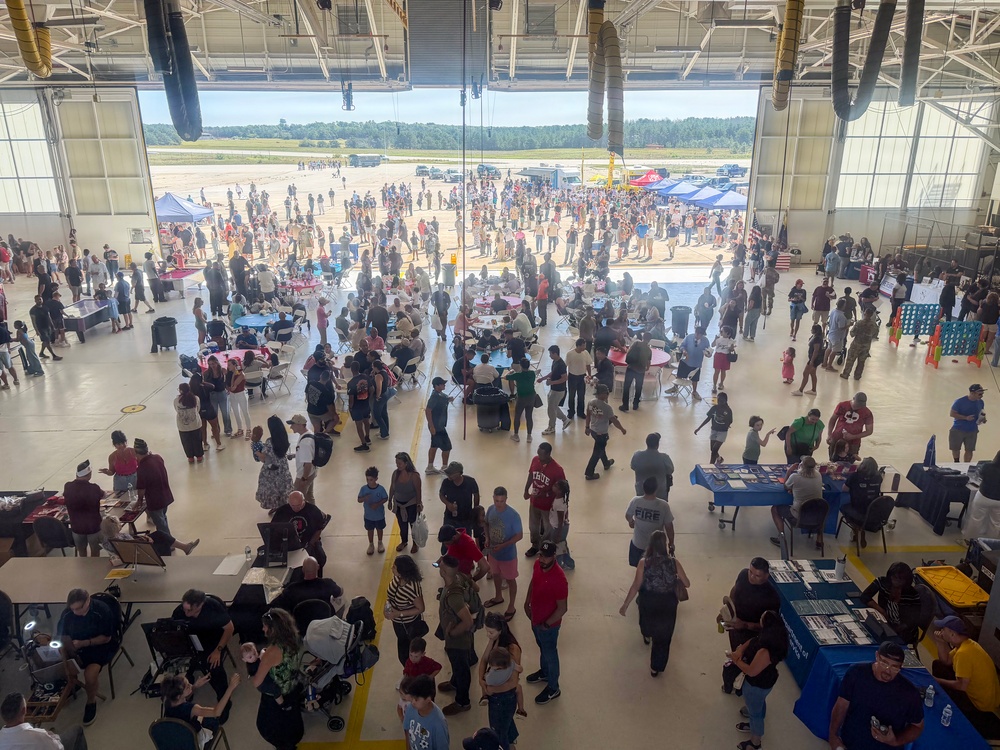A large public event is taking place inside and outside a military or aviation hangar, with hundreds of attendees of all ages milling about. The hangar doors are fully open, revealing an airfield tarmac where additional crowds and vendor tents are visible. The event appears to be a community open house or air show family day with informational booths, games, and outdoor displays.