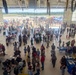 A large public event is taking place inside and outside a military or aviation hangar, with hundreds of attendees of all ages milling about. The hangar doors are fully open, revealing an airfield tarmac where additional crowds and vendor tents are visible. The event appears to be a community open house or air show family day with informational booths, games, and outdoor displays.