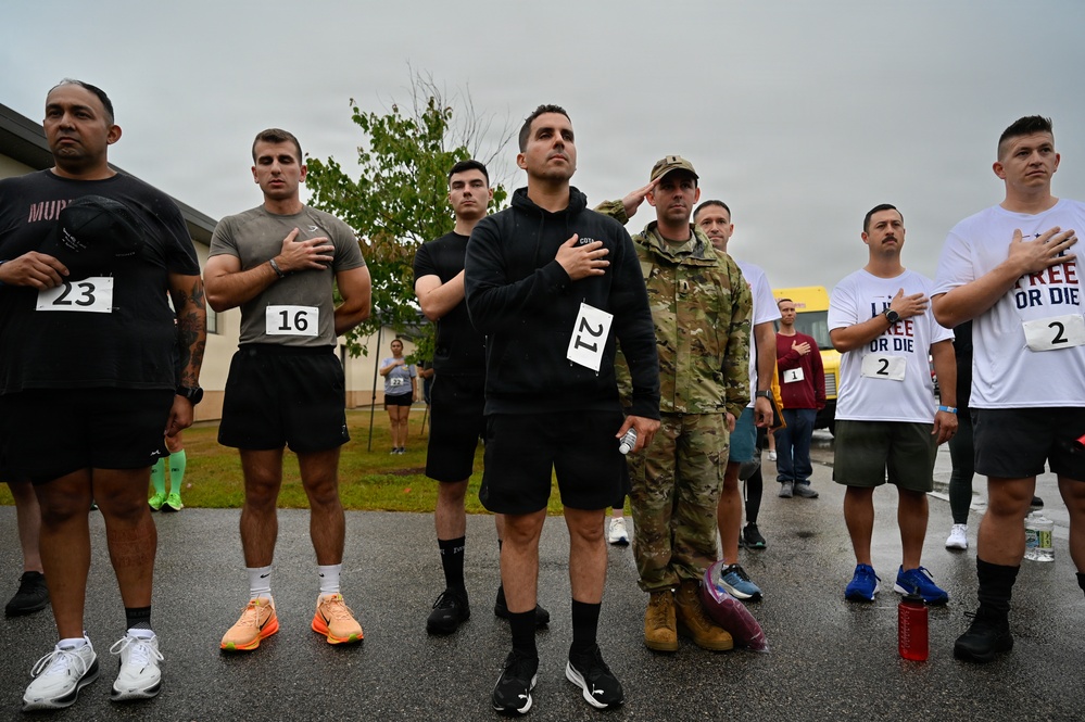 A group of runners and participants stand at attention with hands over their hearts during what appears to be a pre-race ceremony, likely the playing of the national anthem. The event appears to be a military or community-organized road race, with participants wearing numbered bibs (visible numbers include 23, 16, 21, 1, and 2). At least one individual is in military camouflage uniform and is saluting, while others are in civilian athletic wear.