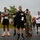 157th Airmen Compete in 2026 Pease Warrior Challenge, Demonstrate Physical Prowess A group of runners and participants stand at attention with hands over their hearts during what appears to be a pre-race ceremony, likely the playing of the national anthem. The event appears to be a military or community-organized road race, with participants wearing numbered bibs (visible numbers include 23, 16, 21, 1, and 2). At least one individual is in military camouflage uniform and is saluting, while others are in civilian athletic wear.