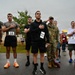 A group of runners and participants stand at attention with hands over their hearts during what appears to be a pre-race ceremony, likely the playing of the national anthem. The event appears to be a military or community-organized road race, with participants wearing numbered bibs (visible numbers include 23, 16, 21, 1, and 2). At least one individual is in military camouflage uniform and is saluting, while others are in civilian athletic wear.