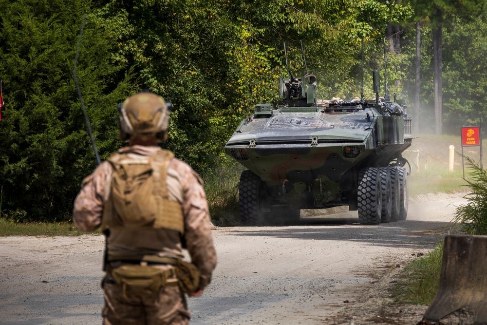 A military training exercise is underway on a dirt road surrounded by dense woodland. A soldier in camouflage uniform with tactical gear and helmet stands facing a large 8-wheeled armored combat vehicle approaching on the road. A red sign marked 'FARM HOUSE' with what appears to be a Marine Corps emblem is visible on the right side of the road.