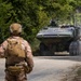 A military training exercise is underway on a dirt road surrounded by dense woodland. A soldier in camouflage uniform with tactical gear and helmet stands facing a large 8-wheeled armored combat vehicle approaching on the road. A red sign marked 'FARM HOUSE' with what appears to be a Marine Corps emblem is visible on the right side of the road.