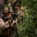 A soldier in desert camouflage (MARPAT pattern) is aiming a rifle through dense green vegetation. The primary subject is in sharp focus in the foreground, while a second individual is visible in the background. The setting appears to be a wooded or forested training or operational environment.