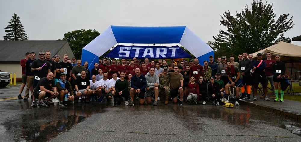 A large group of approximately 60-80 participants poses for a group photo in front of a blue and white inflatable START arch at what appears to be a charity or military fun run event. The wet pavement and overcast sky indicate rainy weather conditions during the event. Participants are wearing race bibs with numbers and a mix of athletic and military-style clothing.