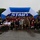 157th Airmen Compete in 2026 Pease Warrior Challenge, Demonstrate Physical Prowess A large group of approximately 60-80 participants poses for a group photo in front of a blue and white inflatable START arch at what appears to be a charity or military fun run event. The wet pavement and overcast sky indicate rainy weather conditions during the event. Participants are wearing race bibs with numbers and a mix of athletic and military-style clothing.