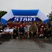 A large group of approximately 60-80 participants poses for a group photo in front of a blue and white inflatable START arch at what appears to be a charity or military fun run event. The wet pavement and overcast sky indicate rainy weather conditions during the event. Participants are wearing race bibs with numbers and a mix of athletic and military-style clothing.