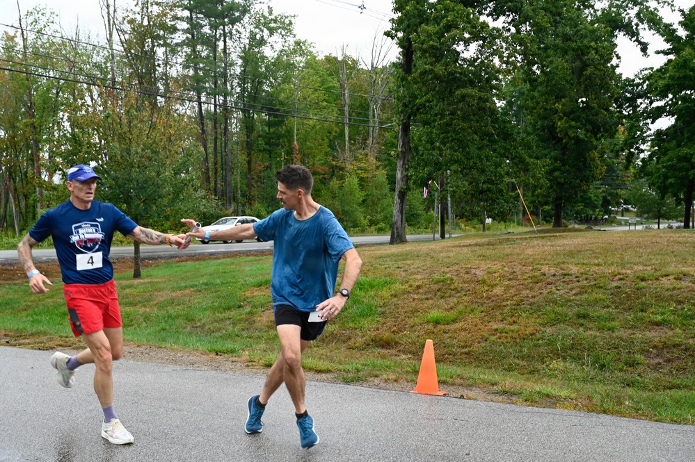 Two male runners are captured mid-stride on a paved road during what appears to be a relay race, with one runner (bib number 4) passing a baton to another runner in a blue shirt. The setting is a rural or suburban area with trees, grass, and a road visible in the background. An orange traffic cone marks the exchange zone on the course.