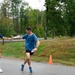 Two male runners are captured mid-stride on a paved road during what appears to be a relay race, with one runner (bib number 4) passing a baton to another runner in a blue shirt. The setting is a rural or suburban area with trees, grass, and a road visible in the background. An orange traffic cone marks the exchange zone on the course.