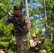 Two military personnel in desert MARPAT camouflage uniforms are conducting what appears to be a tactical training exercise in a wooded environment. The lead soldier is moving forward while carrying a rifle at the ready, while a second soldier in the background maintains a kneeling overwatch position with a scoped rifle. The setting, uniforms, and equipment are consistent with U.S. Marine Corps training operations.