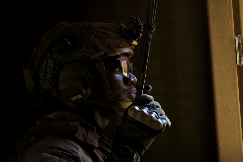 A soldier in full tactical gear is photographed in dramatic low-key lighting, holding a radio communication device to his mouth. He is positioned near what appears to be a doorframe or wall edge, suggesting an indoor tactical environment. The image has a cinematic quality with strong contrast between shadow and light.