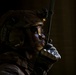 A soldier in full tactical gear is photographed in dramatic low-key lighting, holding a radio communication device to his mouth. He is positioned near what appears to be a doorframe or wall edge, suggesting an indoor tactical environment. The image has a cinematic quality with strong contrast between shadow and light.