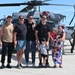 A group of adults and two young children pose for a photo in front of a military helicopter, likely at an air show or open base event. The helicopter appears to be a Sikorsky HH-60 Pave Hawk or similar variant, with tail number 4556 visible. The setting is a tarmac on a clear, sunny day with other visitors visible in the background.