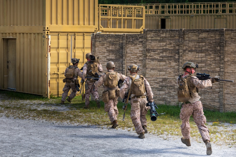 A group of approximately five military personnel in desert camouflage uniforms are conducting what appears to be a tactical training exercise at a mock urban environment. The soldiers are moving in formation along a gravel path toward a yellow shipping container structure adjacent to a stone-textured wall. The lead soldier on the right is actively aiming a rifle with an optic scope while others carry various weapons including what appears to be a rocket launcher.