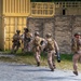 A group of approximately five military personnel in desert camouflage uniforms are conducting what appears to be a tactical training exercise at a mock urban environment. The soldiers are moving in formation along a gravel path toward a yellow shipping container structure adjacent to a stone-textured wall. The lead soldier on the right is actively aiming a rifle with an optic scope while others carry various weapons including what appears to be a rocket launcher.