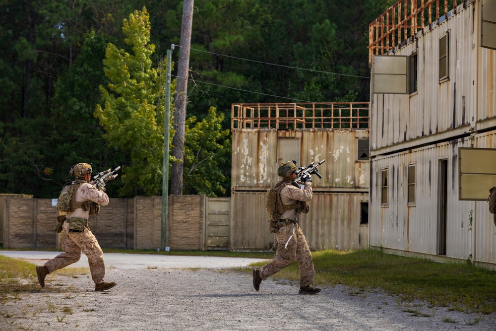Two armed military personnel in desert camouflage uniforms are running through what appears to be a military training facility or mock urban environment. They are carrying assault rifles in a tactical ready position while moving toward a structure made of stacked shipping containers. The setting includes weathered buildings, a perimeter wall, and a wooded background, consistent with a MOUT (Military Operations on Urban Terrain) training site.