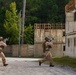 Two armed military personnel in desert camouflage uniforms are running through what appears to be a military training facility or mock urban environment. They are carrying assault rifles in a tactical ready position while moving toward a structure made of stacked shipping containers. The setting includes weathered buildings, a perimeter wall, and a wooded background, consistent with a MOUT (Military Operations on Urban Terrain) training site.