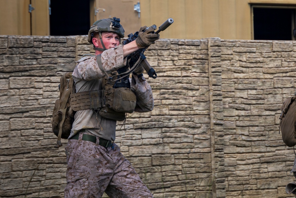 A soldier in desert camouflage (MARPAT pattern) is shown in a tactical stance, aiming a rifle in what appears to be a military training or operational environment. The individual is wearing full combat gear including a plate carrier vest, helmet with night vision mount, and tactical gloves. The background features a stone or concrete block wall structure consistent with a mock urban training facility.