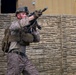 A soldier in desert camouflage (MARPAT pattern) is shown in a tactical stance, aiming a rifle in what appears to be a military training or operational environment. The individual is wearing full combat gear including a plate carrier vest, helmet with night vision mount, and tactical gloves. The background features a stone or concrete block wall structure consistent with a mock urban training facility.