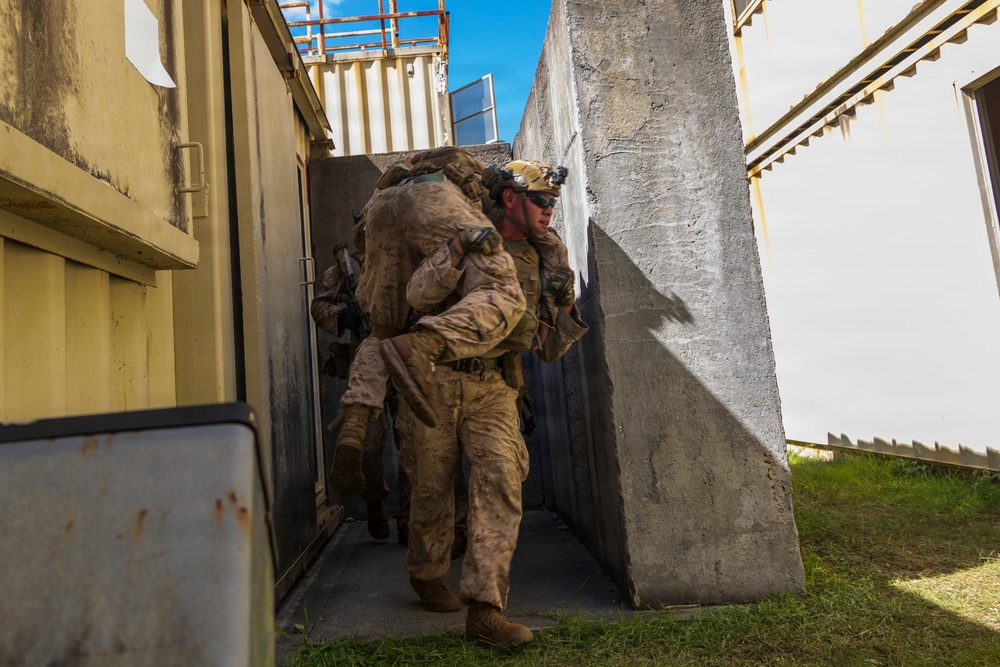 A military service member in desert camouflage (MARPAT pattern) is carrying another person on their back in a fireman's carry through a narrow corridor between concrete barriers and modular/container structures. At least one additional armed service member is visible in the background. The setting appears to be a military training facility or urban operations training environment.
