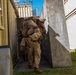 A military service member in desert camouflage (MARPAT pattern) is carrying another person on their back in a fireman's carry through a narrow corridor between concrete barriers and modular/container structures. At least one additional armed service member is visible in the background. The setting appears to be a military training facility or urban operations training environment.