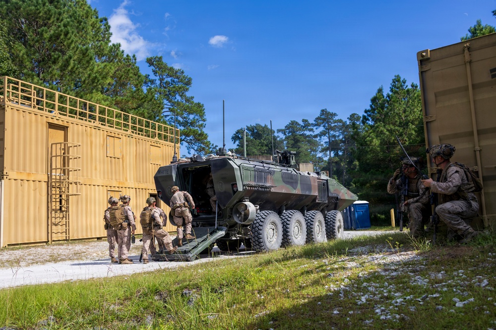 Military personnel in desert camouflage uniforms are conducting a training exercise involving an 8-wheeled armored personnel carrier, likely an ACV (Amphibious Combat Vehicle), positioned between modular container structures. Several soldiers are boarding or dismounting the vehicle while at least one soldier takes a covered position near a tan container wall. The setting appears to be a military training facility with constructed urban terrain elements surrounded by pine trees.