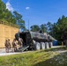 Military personnel in desert camouflage uniforms are conducting a training exercise involving an 8-wheeled armored personnel carrier, likely an ACV (Amphibious Combat Vehicle), positioned between modular container structures. Several soldiers are boarding or dismounting the vehicle while at least one soldier takes a covered position near a tan container wall. The setting appears to be a military training facility with constructed urban terrain elements surrounded by pine trees.