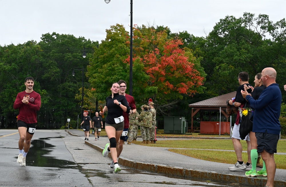 A group of runners wearing race bibs numbered '7' are finishing or participating in an organized running event on a wet road, suggesting recent rainfall. Spectators line the right side of the path, clapping and cheering, while military personnel in camouflage uniforms are visible in the background. The setting appears to be a military installation, with autumn foliage visible in the background trees.