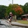 157th Airmen Compete in 2026 Pease Warrior Challenge, Demonstrate Physical Prowess A group of runners wearing race bibs numbered '7' are finishing or participating in an organized running event on a wet road, suggesting recent rainfall. Spectators line the right side of the path, clapping and cheering, while military personnel in camouflage uniforms are visible in the background. The setting appears to be a military installation, with autumn foliage visible in the background trees.