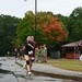 A group of runners wearing race bibs numbered '7' are finishing or participating in an organized running event on a wet road, suggesting recent rainfall. Spectators line the right side of the path, clapping and cheering, while military personnel in camouflage uniforms are visible in the background. The setting appears to be a military installation, with autumn foliage visible in the background trees.