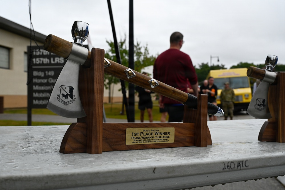 A decorative axe trophy mounted on a wooden stand is displayed in the foreground, awarded for 1st Place Winner of the Pease Warrior Challenge (Male Award). The trophy features a polished metal axe head engraved with a military unit insignia, resting on a walnut-style wooden cradle with a brass nameplate. In the background, several people are gathered outdoors near a building marked '157th LRS' in what appears to be a military base setting.