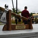 A decorative axe trophy mounted on a wooden stand is displayed in the foreground, awarded for 1st Place Winner of the Pease Warrior Challenge (Male Award). The trophy features a polished metal axe head engraved with a military unit insignia, resting on a walnut-style wooden cradle with a brass nameplate. In the background, several people are gathered outdoors near a building marked '157th LRS' in what appears to be a military base setting.