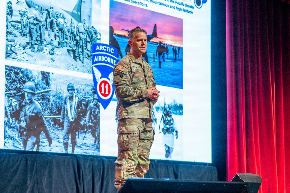 A U.S. Army officer in full camouflage uniform (OCP/MultiCam) stands on a stage delivering a presentation. The backdrop displays a large projection screen showing the '11th Airborne Division - Arctic Airborne' insignia alongside historical and modern military photographs. The setting appears to be a formal military conference or briefing event.